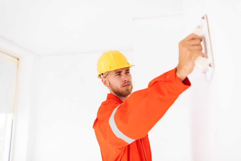 Young Foreman Orange Work Clothes Yellow Hardhat Thoughtfully Using Putty Knife Work Min